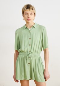 Combinaison courte verte à manches courtes en tissu texturé, avec un devant à boutons, une taille à cordon et deux poches latérales.