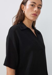 Zwart T-shirt met korte mouwen, kraag en een subtiel textuurpatroon. Bevat een voorzak en heeft een ontspannen pasvorm.