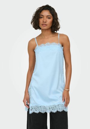 Femme portant un caraco en satin bleu clair avec bordure en dentelle le long de l'encolure, des côtés et de l'ourlet, associé à un pantalon noir et un bracelet à son poignet droit.