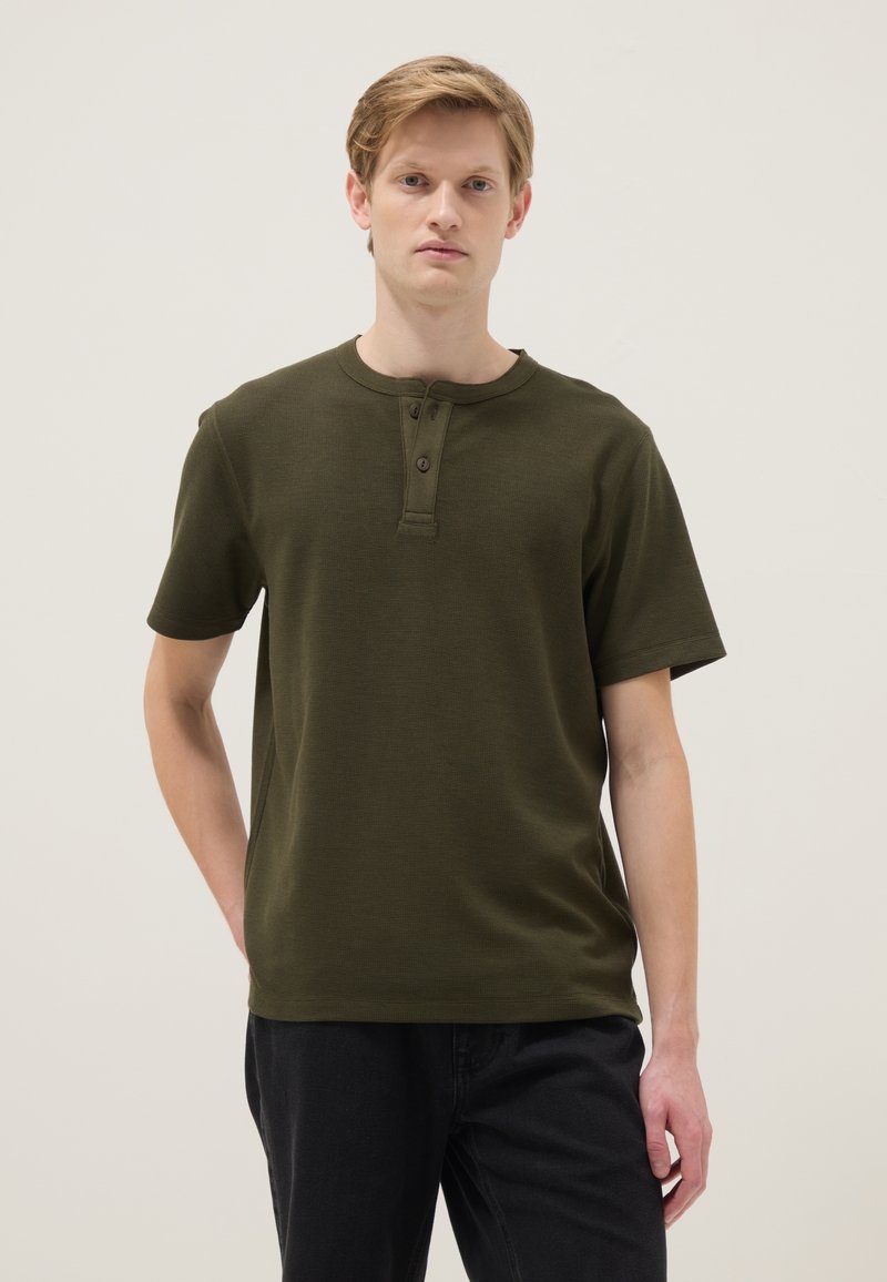 Joven de pie, vistiendo una camiseta henley de manga corta color verde oliva con dos botones y pantalones negros, fondo neutro.