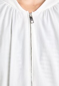 White zip-up garment with a gathered neckline, featuring a silver zipper and smooth, lightweight fabric texture.