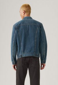 Donkerblauwe denimjack met een kraag, geaccentueerde stiksels op de rug en knoopdetails bij de taille.