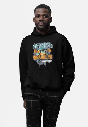 Man draagt een zwarte hoodie met het logo "San Antonio Wreckers Baseball 1997" met een robot die een honkbalknuppel vasthoudt, gecombineerd met een zwarte geruite broek.