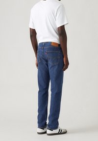 Homme portant un jean bleu, un t-shirt blanc et des baskets blanches avec des rayures noires, debout dos à une surface unie.