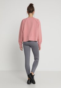 Femme de dos portant un sweat-shirt rose, des leggings gris et des chaussures de sport noires, les cheveux attachés en chignon, sur un fond uni.