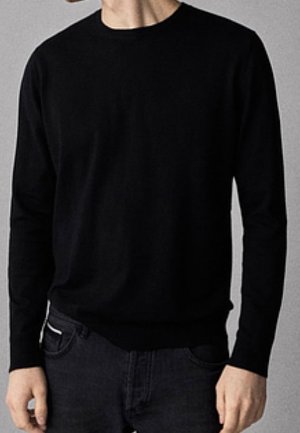 Homme portant un pull noir uni à manches longues et un jean foncé, debout, les mains relâchées le long du corps, sur un fond gris.