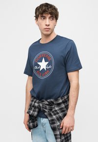 Νέος άνδρας που φοράει μπλε ναυτικό t-shirt Converse All Star και ανοιχτόχρωμο τζιν με μαύρο και άσπρο πουκάμισο καρώ δεμένο γύρω από τη μέση του.