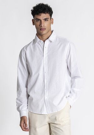 Mand med krøllet mørkt hår iført en hvid skjorte med knapper og beige bukser, står med den ene hånd i lommen mod en ensfarvet baggrund.