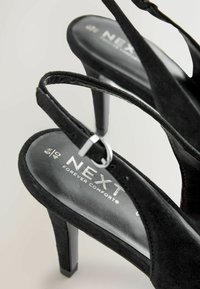 Sandale cu toc din suede negru, cu vârf deschis, curele subțiri și un accent metalic pe toc. Piesa interioară prezintă inscripția brandului.