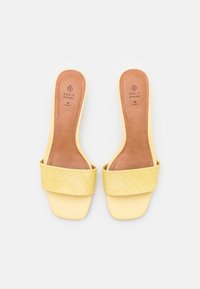 Gule slip-on sandaler med quiltet bred rem og firkantet tå, mærket som veganske, mærket "Call It Spring" synligt på brun indersål.