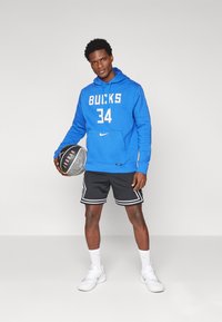 Mėlynas Nike džemperis su užrašu "BUCKS 34", juodos šortai su balta apdaila ir baltos sportinės kedai. Laiko juodą krepšinį. Paprastas fonas.