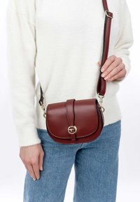 Borsa a tracolla in pelle rosso scuro con superficie liscia, pattina arrotondata, accessori in tonalità oro e tracolla regolabile, indossata all'altezza dei fianchi.