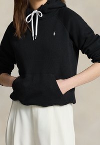 Osoba v černé mikině s bílými šňůrkami a logem, ruce v přední kapse, v kombinaci s volnými bílými kalhotami na neutrálním pozadí.