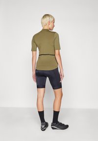 Camisa de manga corta verde oliva y pantalones cortos de ciclismo negros, con un acabado texturizado. Zapatos negros con un detalle de hebilla y calcetines negros.