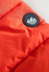 Tissu matelassé orange vif avec un badge rond noir et gris représentant des sommets montagneux et le texte "Adventurer Next Sons 1982."