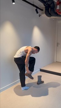 Man in een witte tanktop en zwarte broek bukt zich om een bruine laars aan te trekken in een minimalistische ruimte met witte muren en pijpen aan het plafond.