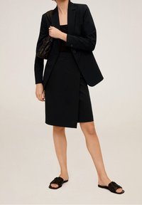Blazer negro entallado combinado con una blusa negra ajustada y una falda envolvente hasta la rodilla. Llevado con sandalias deslizables negras de textura.