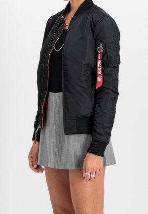 Personne portant une veste bomber noire avec une étiquette rouge « Remove Before Flight » sur la manche, un haut noir et une jupe plissée grise.