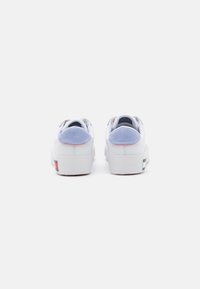 Zapatillas deportivas blancas con una parte superior lisa, acentos de ante lavanda en el talón y una suela de goma texturizada. Presenta una marca multicolor en la parte trasera.