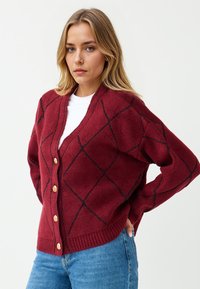 Blondynka ubrana w czerwony kardigan w romby z złotymi guzikami, na białej koszuli i jasnoniebieskich dżinsach, pozująca z rękoma na biodrach.