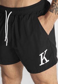 Bañadores negros con cintura ajustable, bolsillos laterales y un logo blanco con la letra "K". Fabricados con un tejido ligero y suave.