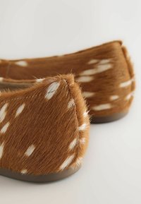 Mocassins en fausse fourrure marron avec des taches blanches et des talons bas sur un fond clair, montrant un gros plan du talon et des détails latéraux.