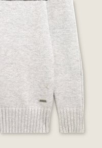 Jersey de punto gris con puños y dobladillo acanalados. Presenta una franja horizontal negra, una etiqueta de logo plateada y una tela suave y texturizada.