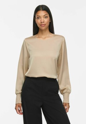 Femme aux longs cheveux foncés porte un chemisier beige à manches longues avec poignets froncés et un pantalon noir taille haute, debout devant un fond blanc.