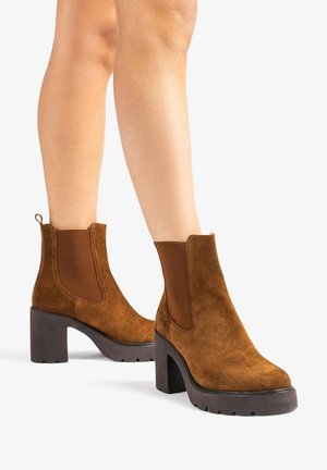 Bruine suède enkellaarsjes met stevige hoge hakken en elastische zijkanten, gedragen op blote benen tegen een witte achtergrond.