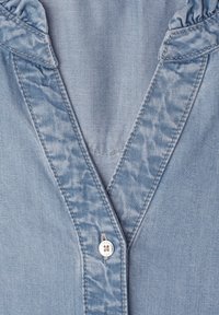 Light blue denim shirt front with V-neckline, ruffled collar, and a single white button with stitching detail.