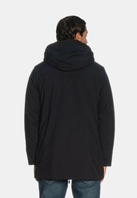 Giacca nera imbottita con grande cappuccio, tessuto testurizzato e forma leggermente allungata, vista di schiena, caratterizzata da un design minimalista.