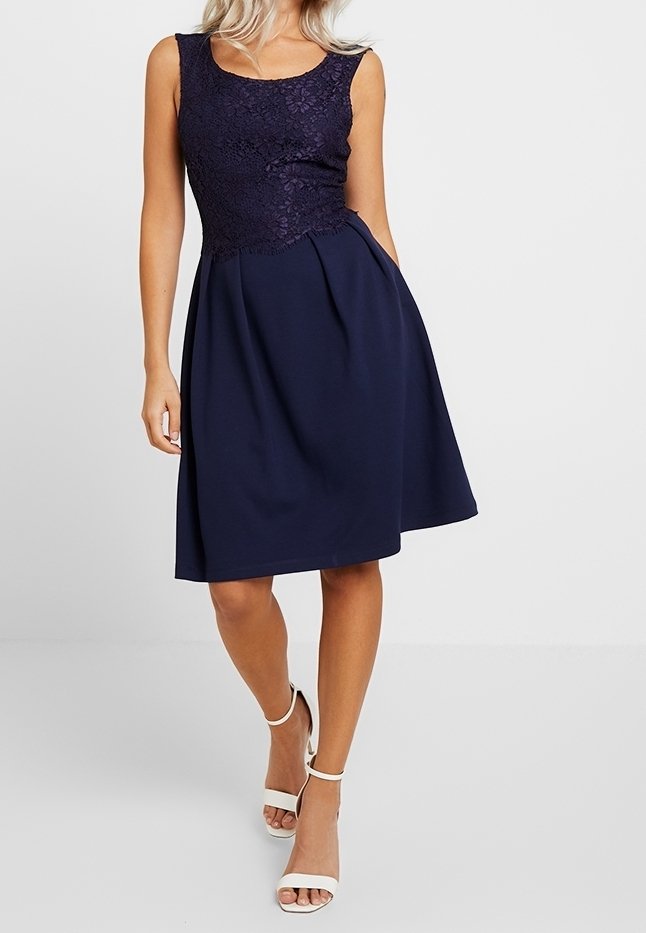 Femme portant une robe bleu marine sans manches avec un corsage en dentelle et une jupe évasée, associée à des sandales blanches à talons hauts avec lanière à la cheville.