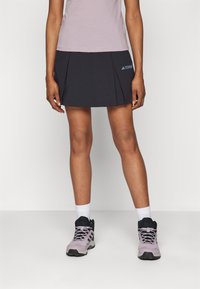 Černý šortkový sukňový kombinace s skládacími prvky a detaily loga, doplněný o světle růžové turistické boty a bílé kotníkové ponožky. Hladká textura a sportovní střih.