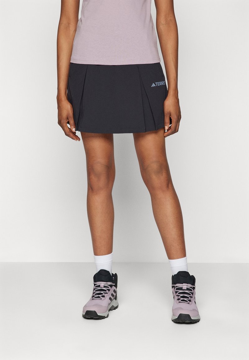 Černý šortkový sukňový kombinace s skládacími prvky a detaily loga, doplněný o světle růžové turistické boty a bílé kotníkové ponožky. Hladká textura a sportovní střih.