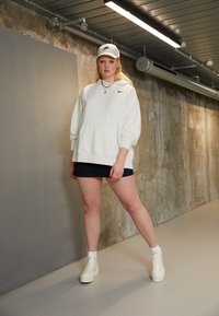 Osoba z długimi blond włosami, nosząca białą czapkę Nike, oversize'owy biały hoodie, ciemną mini spódniczkę i białe buty na platformie, stoi w przemysłowym korytarzu.