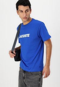 Trumpos rankovės mėlynas medvilninis marškinėliai su balta "LACOSTE" logotipu per krūtinę. Derinami su pilkais džinsais ir juoda petnešų krepšiu.