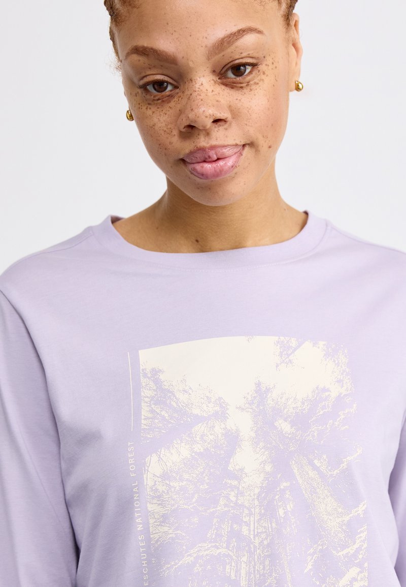 Camisa de manga larga de color púrpura claro con un gráfico sutil de árboles y el texto "Bosque Nacional Deschutes" a lo largo del costado, hecha de una tela suave y lisa.