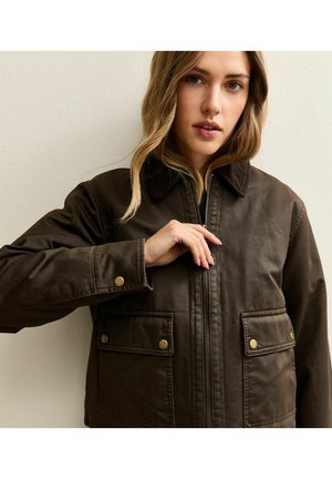 Femme aux cheveux châtain clair portant une veste marron foncé avec une fermeture éclair et des poches avant à boutons, ajustant la fermeture éclair.