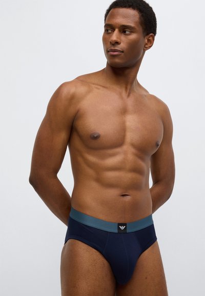 Tamsiai mėlynos spalvos trumpikės iš lygaus audinio, su kontrastingu žalsvu elastiniu juosmeniu ir juoda logotipo etikete. Aptempto prigludimo, ergonomiškas dizainas.