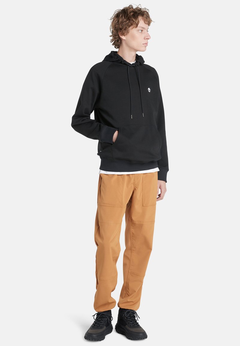 Schwarzer Hoodie mit einer vorderen Tasche und verstellbarem Kordelzug, kombiniert mit lockeren, hellbraunen Cargo-Hosen mit aufgesetzten Taschen und schwarzen Sneakers.