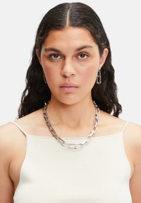 Sølvmetal kædedesign med rektangulære led, parret med matchende rektangulære øreringe. Modellen bærer en lys tanktop.