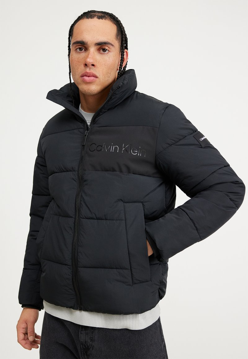 Juodas pūkinis džemperis, pagamintas iš nailono, su užtrauktuku, aukštu apykakle ir priekinėmis kišenėmis; su įspauduotu "Calvin Klein" logotipu.