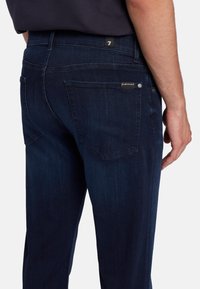 Jeans in denim blu scuro con una vestibilità slim, dotati di due tasche posteriori e di una piccola etichetta con logo. Texture morbida con dettagli di cucitura visibili.