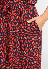 Námořnicky modré šaty s červeným a broskvovým listovým vzorem, elastickým pasem a kapsami. Hladká textura s volným střihem. Doplňky v barvě zlata.