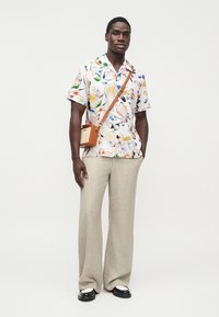 Tričková košeľa s krátkym rukávom s farebným kvetinovým vzorom, béžové široké nohavice a malá hnedá kabelka cez rameno. Čierno-biele dvojfarebné topánky.