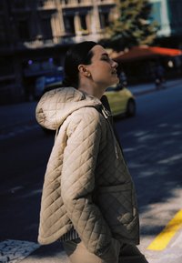 Mladi oseba v bež prešiti jakni s kapuco, stoji na mestni ulici na prostem, z zaprtimi očmi in sproščenim izrazom.