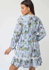 Mujer vestida con un vestido floral azul de mangas largas, con bordes de encaje y dobladillo con volantes, mirando por encima de su hombro contra un fondo liso.