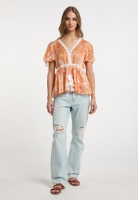 Blusa floreale arancione con bordo in pizzo bianco e dettagli con frange, abbinata a jeans chiari strappati e sandali marroni a ciabatta.