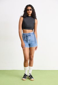 Zwarte mouwloze crop top met hoge hals, gecombineerd met lichtblauwe denim shorts met een strik taille. Model draagt zwarte sneakers en witte sokken.