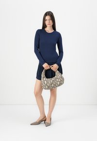 Kvinne står med en beige veske med slangeskinnmønster, iført en marineblå langermet topp, plyndret skjørt og taupe slingback kitten hæler.